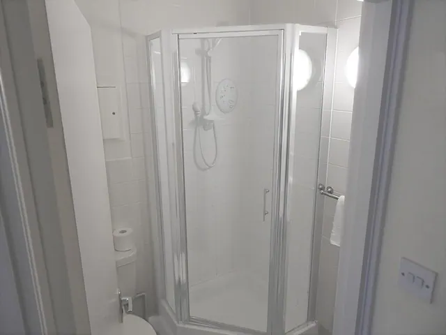 A white tiled corner shower cubicle with glass doors and an electric shower unit, with a toilet partly visible at the left edge.