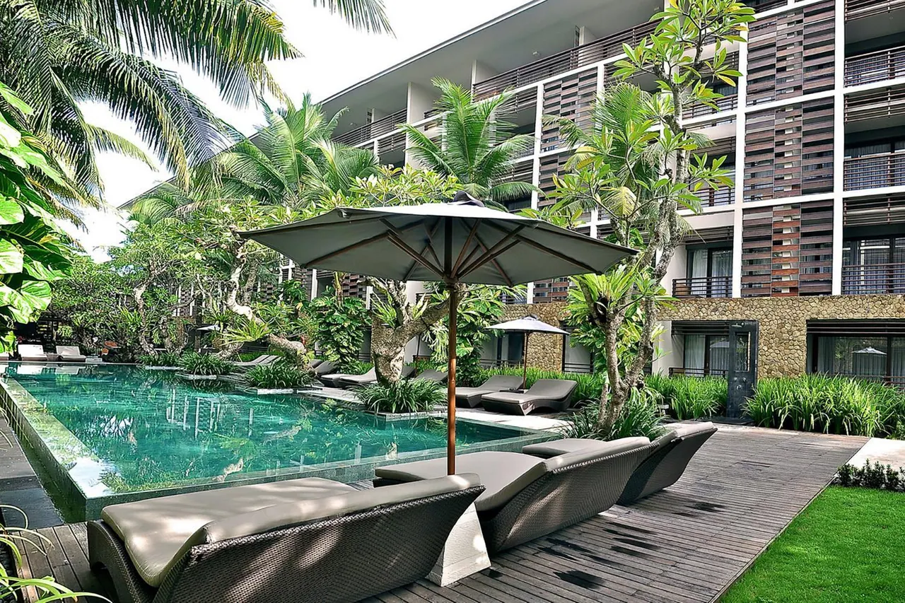 A green-tiled pool with grey wicker loungers and a parasol beside it, with a multi-storey hotel building and palm trees behind.