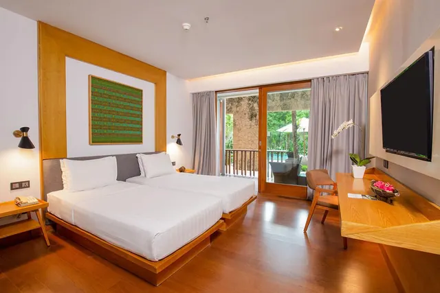 A hotel bedroom with two beds, a wall-mounted television and a desk, with sliding doors open onto a balcony overlooking a pool and trees.