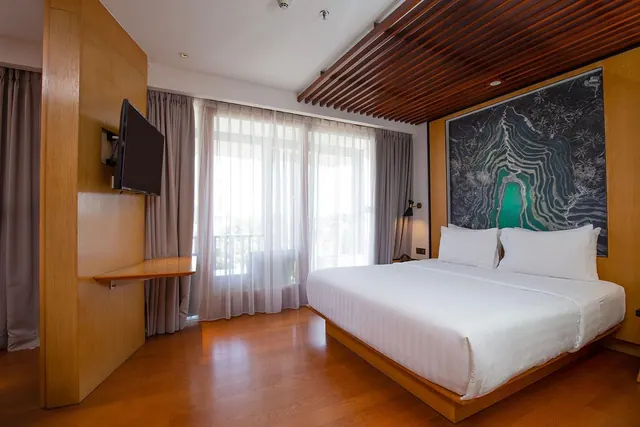 A bedroom with a king bed under a dark wall artwork, a wall-mounted television, and sheer-curtained windows leading to a balcony.