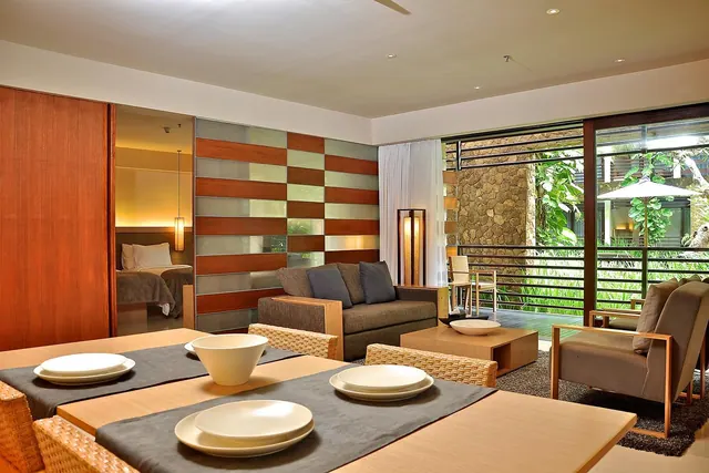 A living area with a dining table in the foreground, a sofa and armchairs, and sliding glass doors opening onto a terrace with greenery outside, with a bed visible through a glass partition on the left.