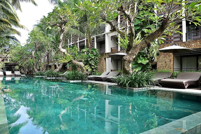 A turquoise swimming pool with sun loungers and white parasols sits in front of a multi-storey building framed by tropical trees.