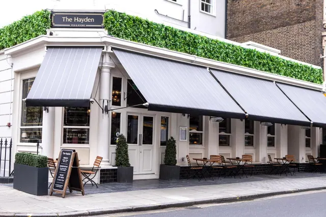 The exterior of The Hayden pub, with grey striped awnings, green planting along the roof, and outdoor tables and chairs on the pavement.