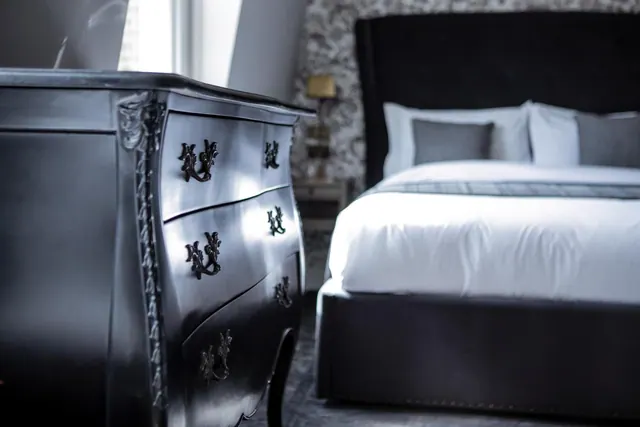 A black bombé chest of drawers stands in the foreground beside a bed with a black upholstered headboard and white bedding.
