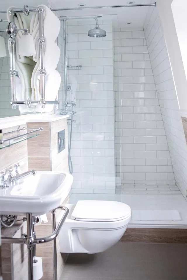 A white bathroom with a walk-in tiled shower behind a glass screen, a wall-hung toilet, a pedestal basin with chrome legs, and a towel rail holding white towels.