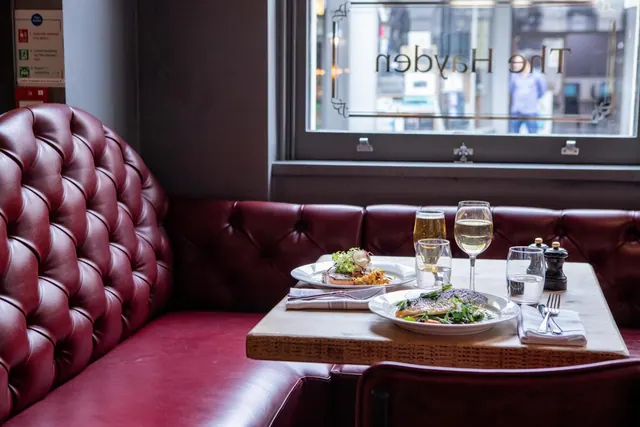 A restaurant booth with red tufted leather seating and a table set with two plated dishes, drinks and cutlery, with a window showing the street outside.
