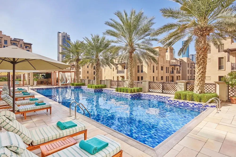 The pool is lined with sun loungers with teal towels and a beige umbrella, with palm trees and beige apartment buildings behind.