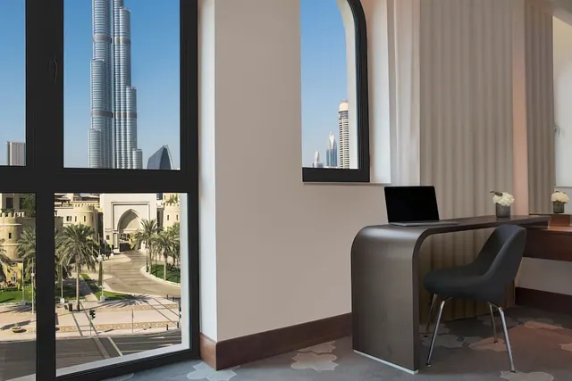 A desk with a laptop and grey chair stands by a window framing the Burj Khalifa and a street lined with palm trees below.