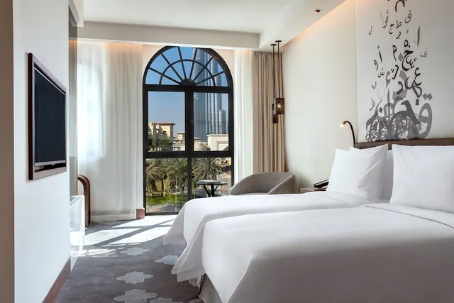 A hotel bedroom with two white-made beds and an arched window looking out at the Burj Khalifa and nearby palm trees.