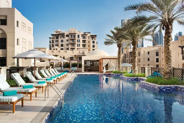 An outdoor swimming pool with blue mosaic tiles is lined with sun loungers under white umbrellas and palm trees, with a cabana and residential buildings in the background.