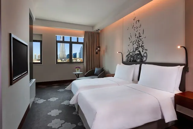A twin-bedded hotel room with a wall-mounted television, an armchair by the window, and a view of the Burj Khalifa and surrounding towers.