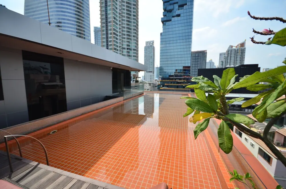 A rooftop swimming pool with orange tiles, surrounded by high-rise towers, with a frangipani plant in the foreground.