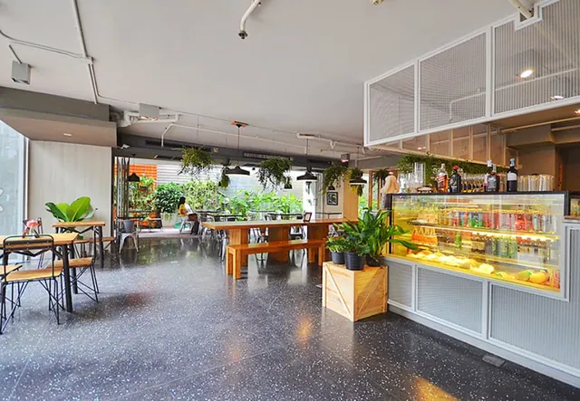 A café dining area with wooden tables and benches on a terrazzo floor, with a drinks display fridge at the bar counter on the right.