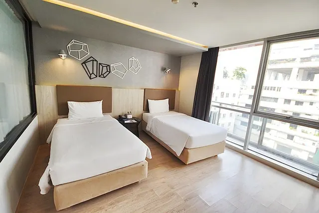 Two single beds with white bedding stand in a bedroom with floor-to-ceiling windows overlooking nearby buildings.