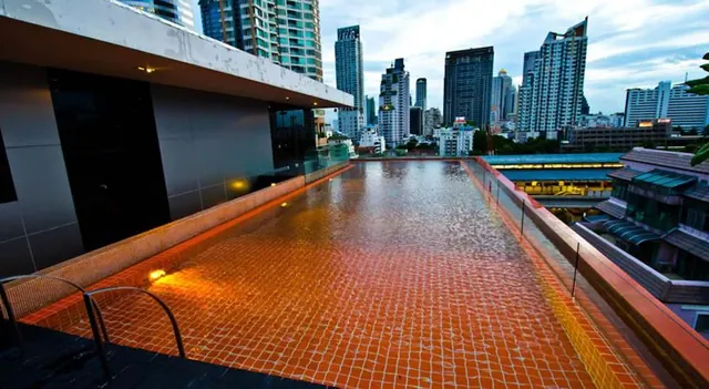 A rooftop swimming pool with an orange-tiled floor and lit edges looks out over a city skyline of high-rise towers at dusk.