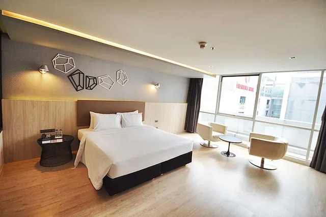 A bedroom with a made king bed, armchairs beside a small round table, and floor-to-ceiling windows looking out onto neighbouring buildings.