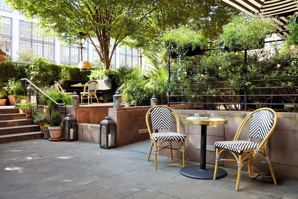 A paved outdoor terrace with bistro chairs, a round gold-topped table, potted plants, lanterns and a striped umbrella, with a large building behind the greenery.