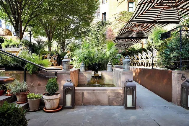 An outdoor courtyard garden with a stone water feature, potted plants, lanterns and striped umbrellas over chairs.