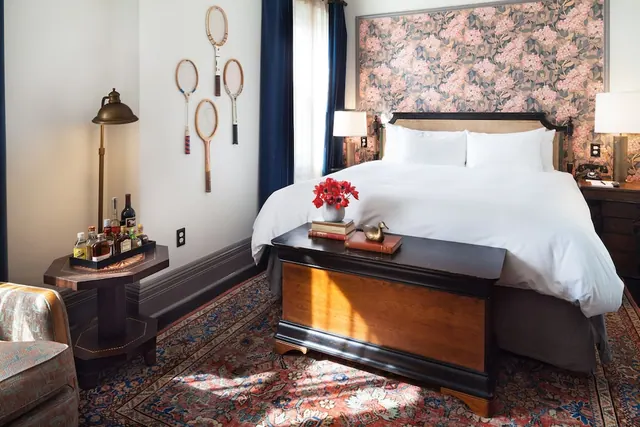 A bedroom with a made king bed, floral wallpaper behind the headboard, a wooden chest at the foot of the bed, and tennis rackets mounted on the wall.