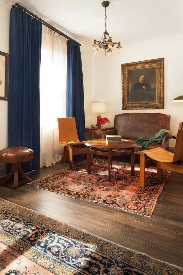 A furnished sitting room with a leather sofa, wooden armchairs, a round coffee table, layered rugs, blue curtains and a framed portrait.