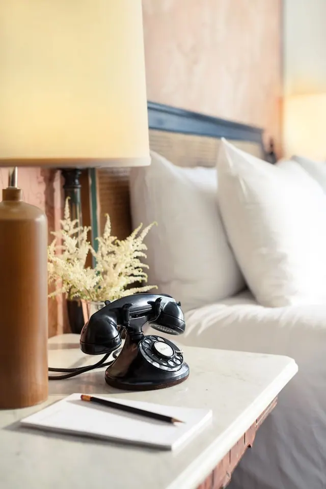 A black vintage rotary telephone, a notepad and a pencil sit on a marble bedside table beside a lamp and a made bed with white pillows.
