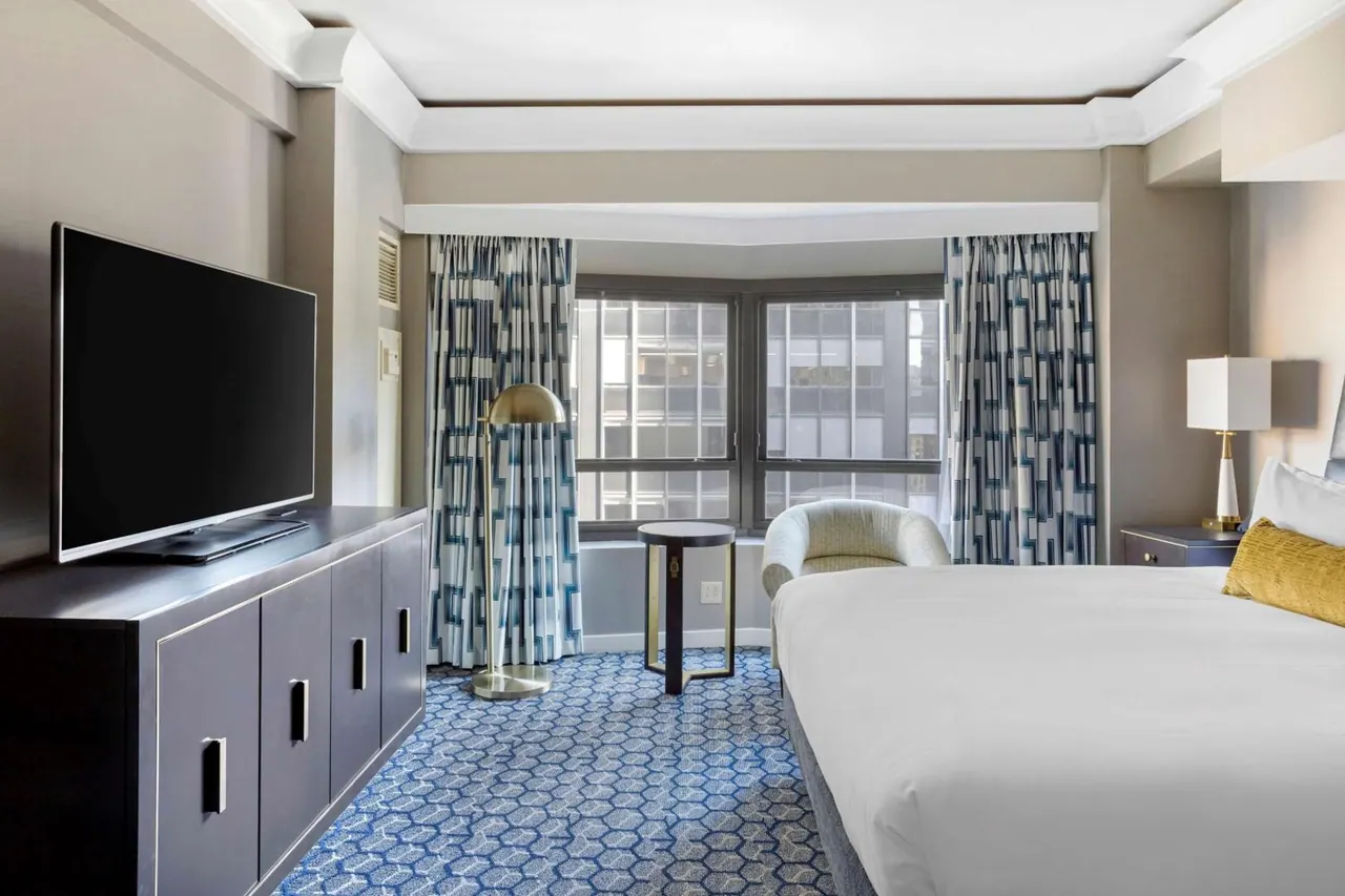 A bedroom with a made king bed, a wall-mounted television on a dark credenza, a armchair and floor lamp, and a bay window looking out onto neighbouring buildings.