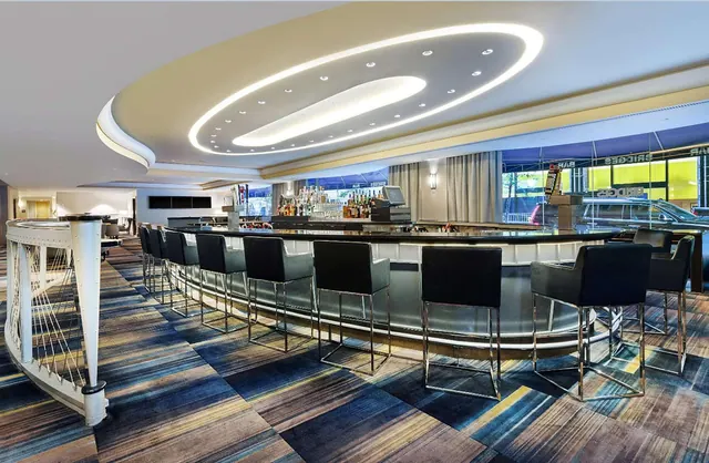 A curved bar with black upholstered stools sits under an oval ceiling light, with windows onto a street and a parked car outside.