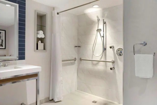 A white tiled bathroom with a walk-in shower fitted with grab rails and a handheld showerhead, next to a vanity sink, with a folded towel on a wall ring.