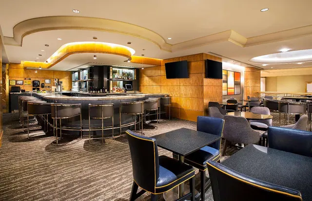A hotel bar and lounge with a curved bar counter, bar stools, dark tables and upholstered chairs, and wall-mounted televisions.