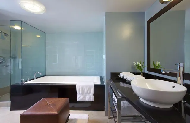 A bathroom with a built-in bathtub beside a glass-walled shower, a vessel sink on a black vanity, and a brown leather ottoman on the floor.