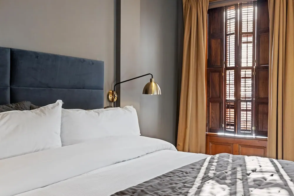 A bedroom with a made bed, a navy upholstered headboard, a brass wall lamp, and a window with wooden shutters and beige curtains.