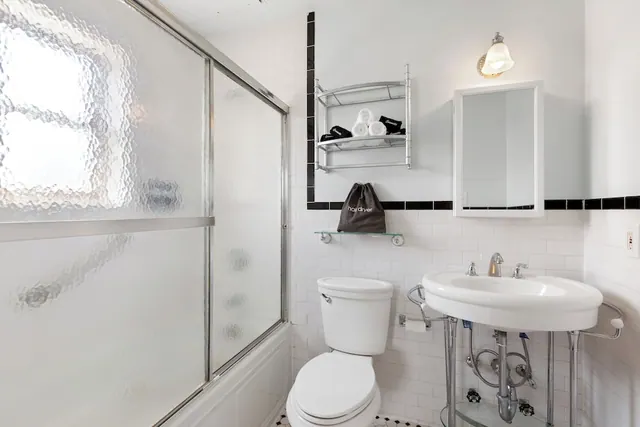 A white bathroom with a glass-enclosed shower over a tub, a pedestal sink, a toilet, and a wall shelf holding rolled towels.