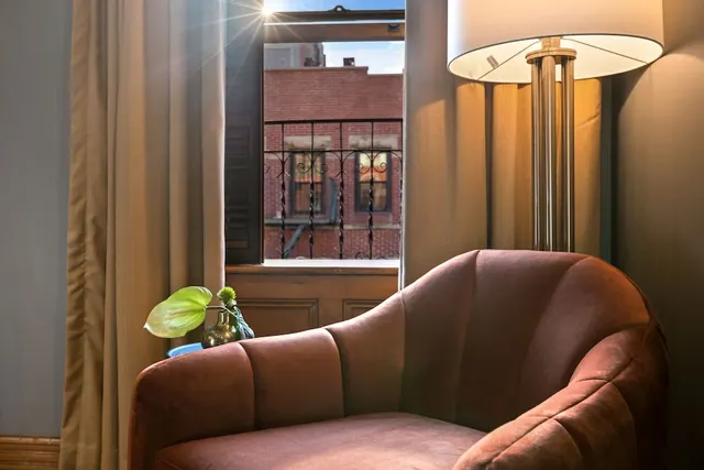 An upholstered armchair sits beside a floor lamp and a window that looks onto a brick building across the way, with a plant in a vase on a small side table.