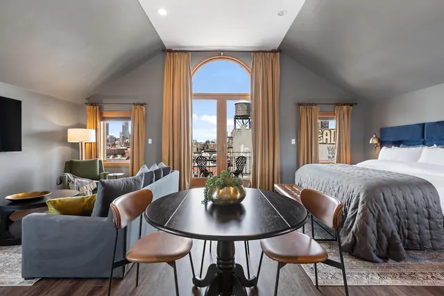 A hotel room with a king bed, a sofa and armchair seating area, a round dining table, and arched French doors opening onto a balcony with a view of city buildings and a rooftop water tower.