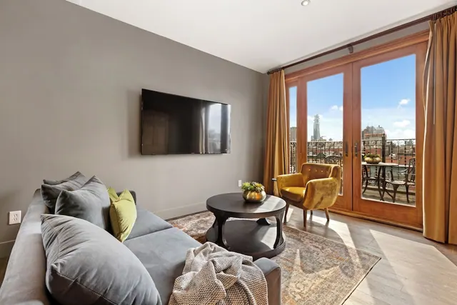 A furnished living room with a wall-mounted television, grey sofa, round coffee table and yellow armchair, with glass doors opening onto a balcony with a city view.