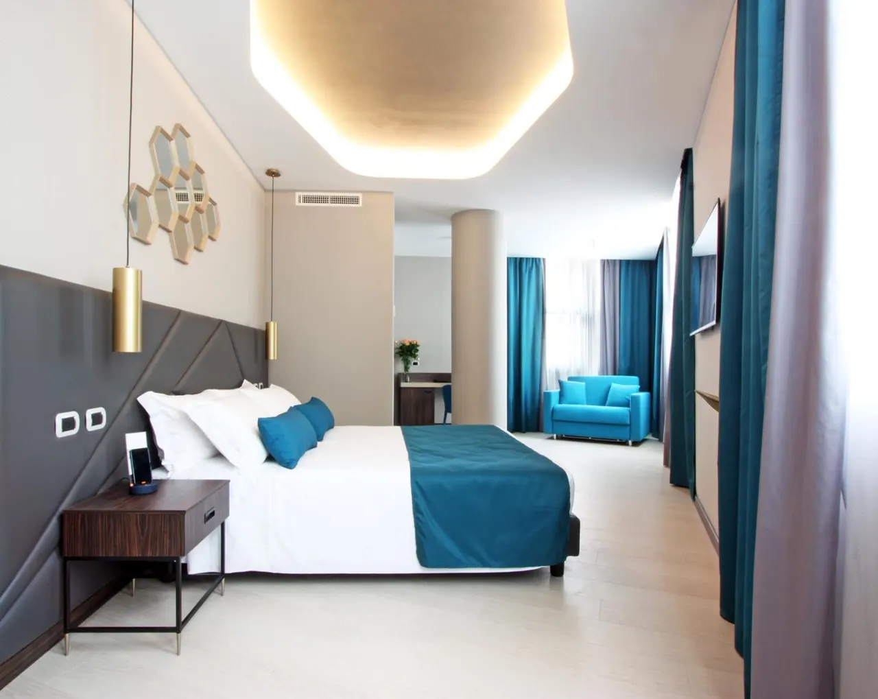 A bedroom with a made bed with a teal throw, a blue armchair by the window, a wall-mounted television and a desk in the background.