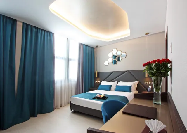 A bedroom with a double bed dressed in teal and white bedding, teal floor-length curtains, a wall of hexagonal mirrors and a desk holding a vase of red roses.