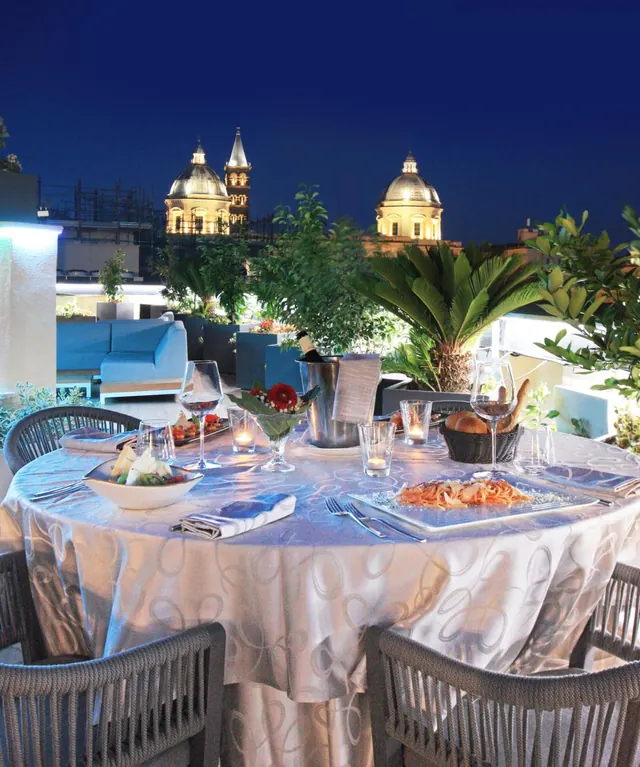 An outdoor rooftop dining table is set with linen, glasses, bread and a wine bucket, with illuminated church domes visible at night behind it.
