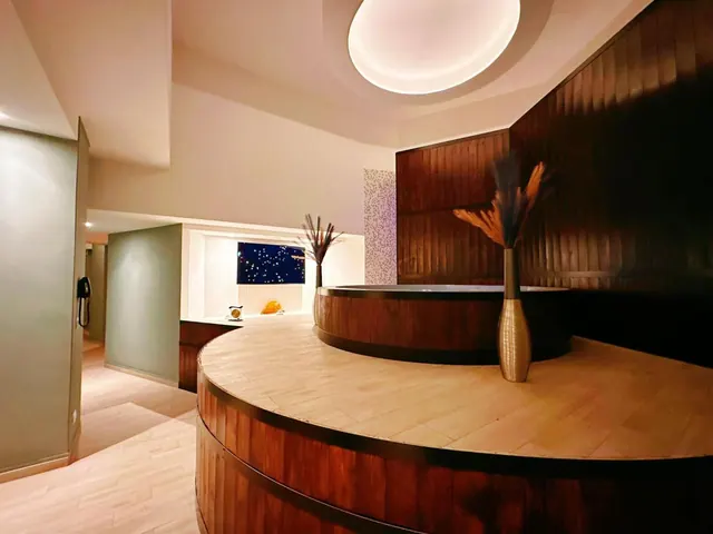 A curved wooden reception counter holds two tall vases of pampas grass beneath a glowing round ceiling light, with a backlit starry blue panel behind it.