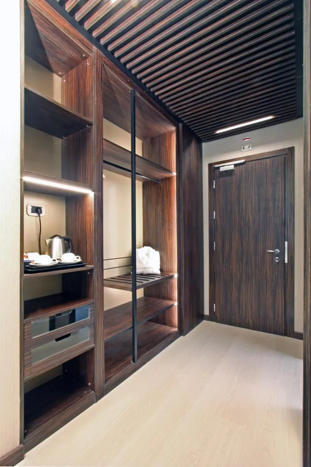A slatted wooden ceiling and dark wood-panelled wardrobe with open shelves holding a kettle and cups stand beside a wooden room door.