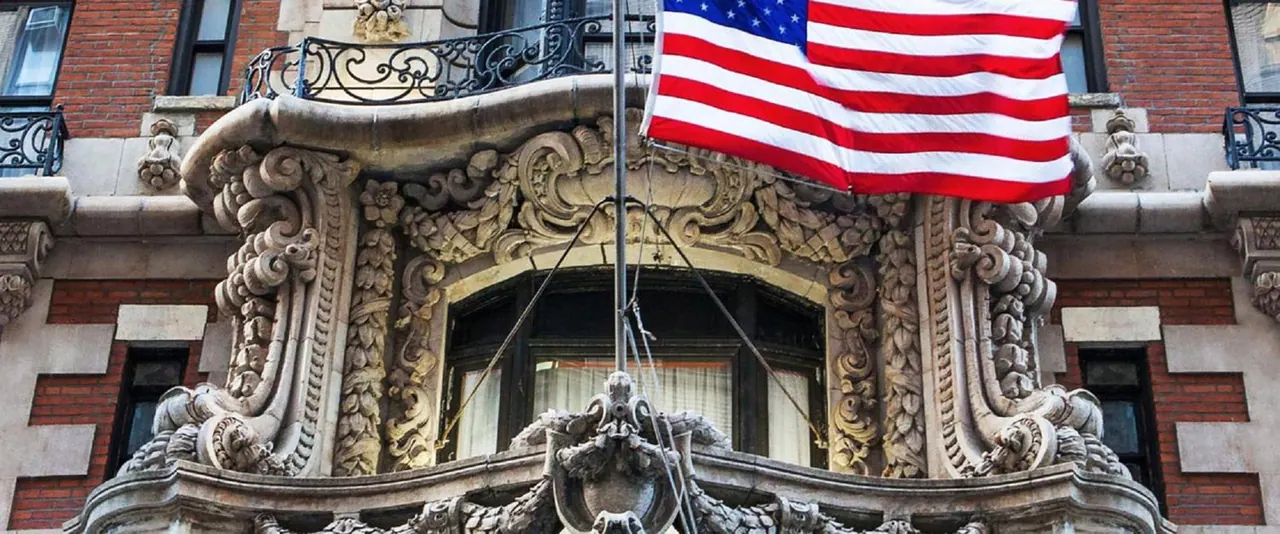 The ornate carved stone bay window and facade of a brick building, with an American flag flying from a pole above.