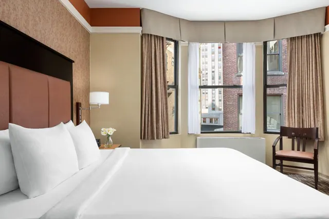 A made king bed with white linens sits in a room with a wooden armchair by the window, which looks out onto nearby brick and high-rise buildings.