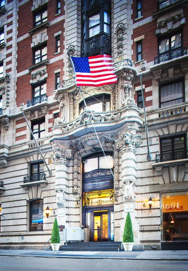 The ornate stone and red brick facade of a building with an American flag, an arched entrance flanked by two topiary planters, and an illuminated storefront at right.