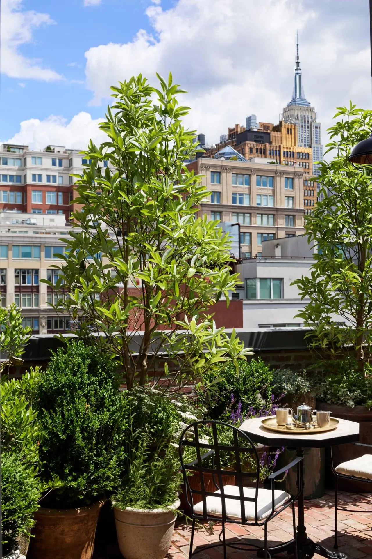 A rooftop terrace with potted trees, a small round table set with a tea service and wrought iron chairs, with the Empire State Building and surrounding buildings visible behind.