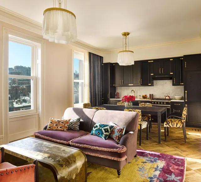 A living area with a plum velvet sofa, a dining table and a dark kitchen, with tall windows looking out over city rooftops and buildings.