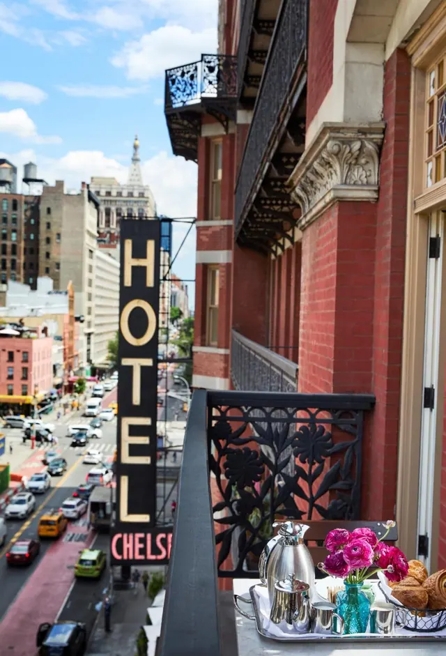 A breakfast tray with a silver pot, pink flowers and croissants sits on a wrought iron balcony overlooking a city street and a vertical Hotel Chelsea sign.