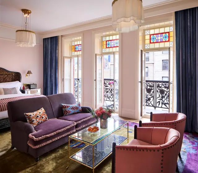 A bedroom with a bed on the left, a purple velvet sofa, pink armchairs, and open French doors onto a balcony with ornate ironwork railings.