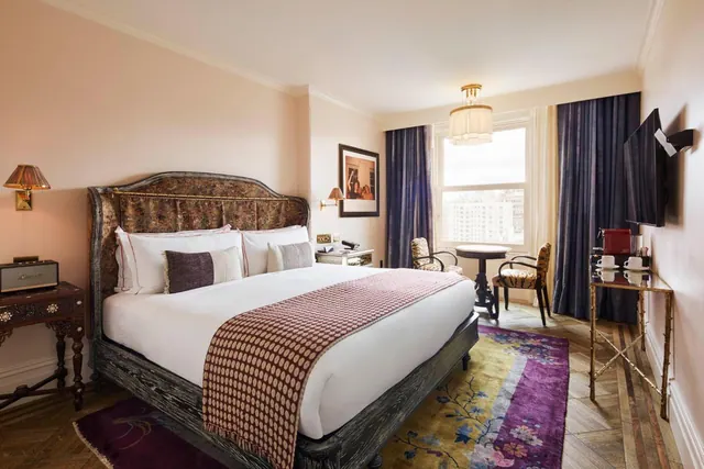 A bedroom with a king bed under an upholstered headboard, two armchairs by a window overlooking buildings, and a wall-mounted television.