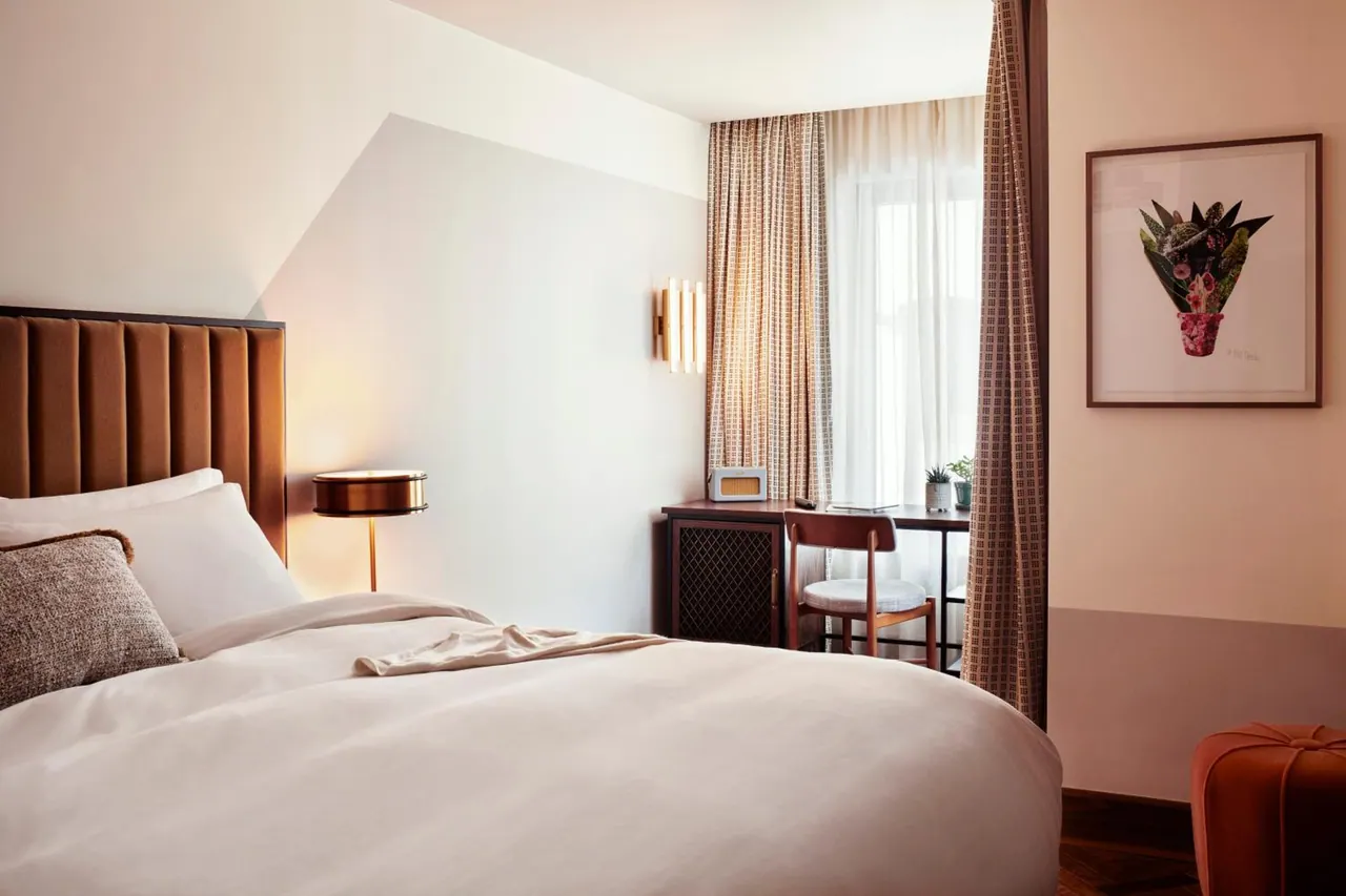 A bedroom with a made double bed, a bedside lamp, a desk and chair beneath a window, and a framed botanical print on the wall.