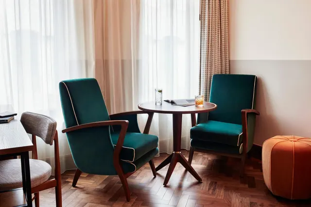 Two teal armchairs sit either side of a small round wooden table holding a drinking glass and a open book, beside sheer curtains and a parquet floor.